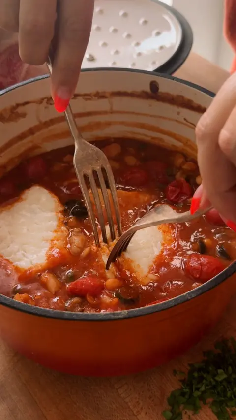 A orange pot full of white fish and cannellini bean puttanesca with two forks inside