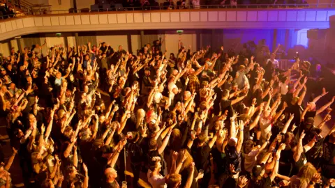 A large crowd of people stood in front of the Town Sall stage with their arms pointing upwards 