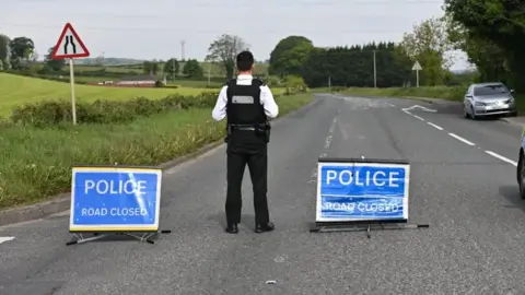 Pacemaker Police officer facing away from camera standing in the middle of the road with two signs that read 