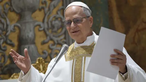 Pope Leo, in white and gold robes, standing at a microphone in the Sistene Chapel. He holds a piece of paper. There is gold and grey artwork on the wall behind him.