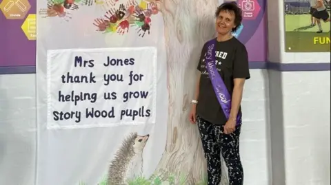 Joanne Jones Joanne is stood by a poster hanging from a wall at a school. She is wearing dark leggings and a T'shirt with a purple sash across her front. The poster is from pupils at Story Wood School and thanks her for helping them.