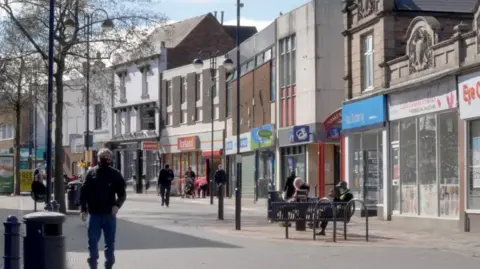 Bilston High Street has shops on both sides with people walking along it and sat on a bench.