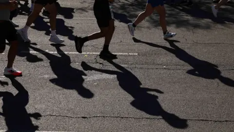 View of the legs of about half a dozen runners on a road. They are in silhouette, casting shadows on the tarmac surface, which has a crack running down it towards the bottom of the image.