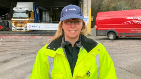 Renee O'Mahony - a blonde woman in high-vis jacket and blue baseball cap in front of a warehouse