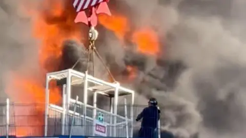 Chris Lauder/PA Media Flames surround a cage being lifted by a crane. A man is getting inside.
