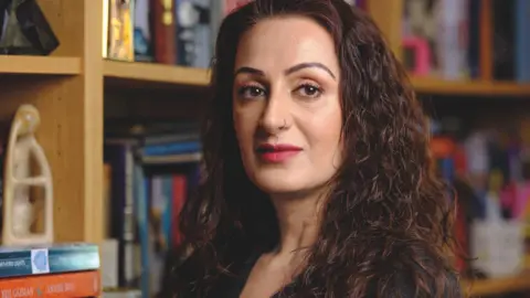 Syima Aslam, a woman of south Asian heritage with curly dark hair standing in front of a book case. She is wearing lipstick and has a nose piercing. 