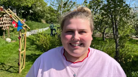Ollie Conopo/BBC A woman in a pink T-shirt and silver necklace stands in a garden with a wooden structure behind her.