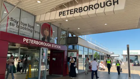 Emma Baugh/BBC The entrance to Peterborough Station with a large sign above the entrance saying Peterborough Station and men and women walking along the concourse and exciting the station