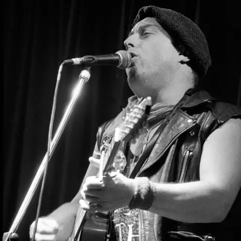 Getty Images A black and white photo of a man singing into a microphone while playing a guitar in front of a black backdrop. He is wearing a black flat cap and a sleeveless leather jacket.