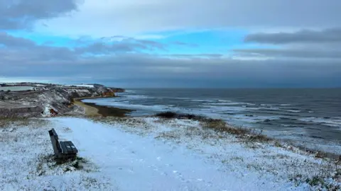Jill Reed A bench at the top of snow-covered cliffs overlooks the sea waves and the dark blue sky. A beach is visible in the distance.