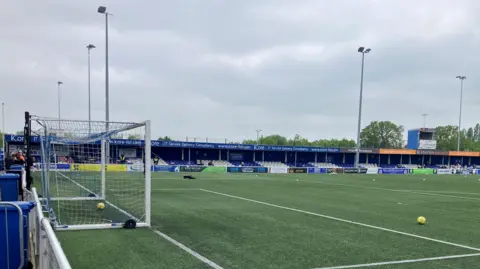 Warren Muggleton/BBC A green artificial football pitch, with a goal to the lefthand side, and a yellow football sitting on the pitch to the right. In the background is blue and white-painted seats in a stand. There are floodlight pillars towering above. It is cloudy.