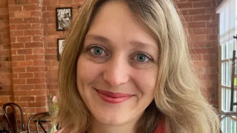 Tetiana Hornik smiling at the camera standing in front of a brick wall, with long blonde hair. Pictures are on the wall behind her. 