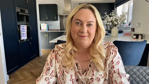 BBC / Natalie Bell Chasing Rainbows founder Sam Catanach. She has blond shoulder-length hair and is wearing a light-coloured patterned shirt. She is sitting in her kitchen.