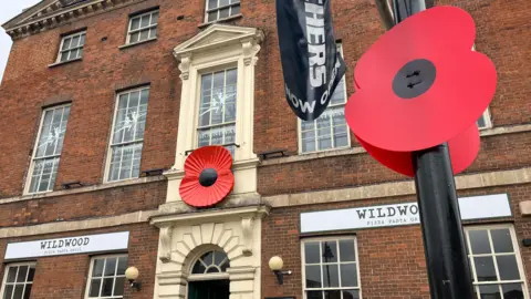 A red poppy is hung in the front of a red-brick building. To the right of the picture frame is a paper poppy hung on a black lamp post.