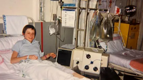 Maddy Warren A girl lies in a hospital bed next to a dialysis machine. She smiles at the camera.
