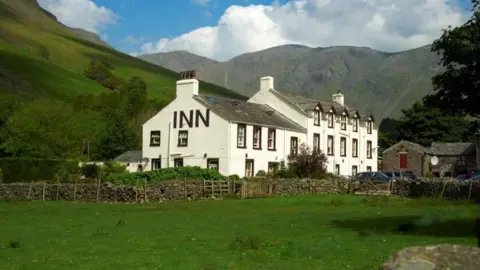A general view of the Wasdale Head Inn. It is a traditional Lakes building, painted white with black detailing around the sash windows. and a slate pitched roof. It is set within a remote setting, with the mountains behind it and a grassy field in front of it