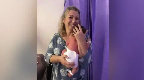 Verina Henchy holding a baby and smiling at the camera, with purple curtains in the background