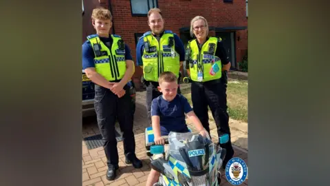 A young boy sitting on a toy police motorbike with three police officers standing behind him. They are wearing blue t-shirts and fluorescent yellow police vests