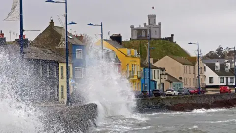 An image of the seafront at Donaghadee, colourful houses can be seen along the promenade while waves break against the sea wall and spray up onto the road. It is an overcast day.