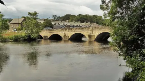 Geograph The existing bridge over the Wharfe in Otley is a seven-span stone bridge which dates back to the 13th Century.  It is shown here with houses in the background and trees on the river bank.