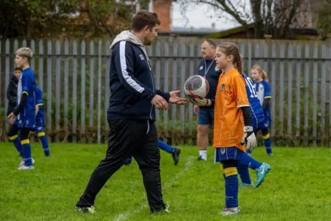 Thomas Meacham A male football coach and a young girl are in focus with more kids, male and female, in the background, as well as another male coach. They are all kitted up and the young girl is handing a ball over to the coach. In the background there is a grey fence and houses behind it.