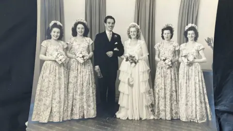 Supplied The image is a black-and-white studio photograph. There are seven people standing side by side, facing the camera. The arrangement is symmetrical: One woman stands at the centre slightly forward, wearing a layered, floor-length light-coloured outfit. The man immediately to the left of the central figure wears a dark suit with a tie. The remaining five people wear similar long dresses with matching patterns. All individuals are standing upright with composed posture, holding floral bouquets except for the person in the dark suit.