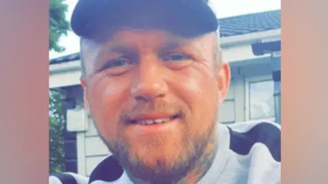 A photo of Marcus Carpenter. He is smiling at the camera, he has a beard and tattoos on his neck. He is wearing a grey sweatshirt with black details, and a black cap.