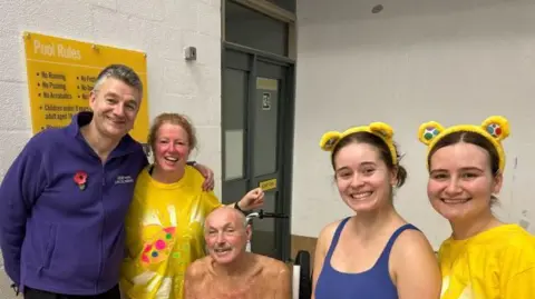 Steve Harrison is sat down smiling with members of the Lincoln BBC swim team. Two women are wearing Pudsey ears, another woman is wearing a Pudsey yellow t-shirt and a man is wearing a purple BBC Radio Lincolnshire fleece with a Remembrance poppy attached to it.