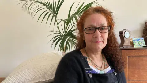 Elizabeth Donowho in a living room. There is a plant behind her and she is wearing glasses. 