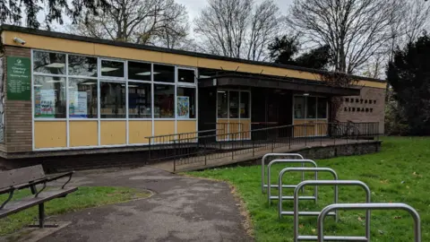 Google The outside of Chertsey Library, showing the sign on the door, with a park bench in front of the building, along with bicycle racks.