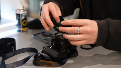 A close-up view of hands assembling a camera on a countertop. The person is attaching a lens cap to a black digital camera, which has a strap resting on the surface. Nearby, there is a lens hood, a smartphone, and a canister of cleaning spray in the background. 
