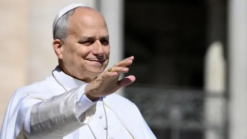 EPA Pope Leo XIV, wearing all white, waving his right hand in the air. 