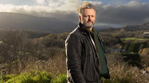 Standing on high ground above a valley, Michael Sheen looks towards the camera with rolling hills and patchwork fields stretching into the distance. He is wearing a brown jacket with a green patterned jumper. Sheen has grey hair and facial hair. Strong afternoon sunlight and heavy clouds create dramatic contrasts across the rural landscape.