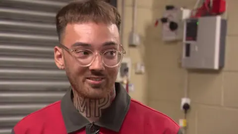 Max, a man with brown hair, clear glasses and tattoos, in a red and grey Iceland uniform speaking to the BBC from inside a warehouse