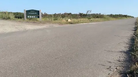 A quiet junction in a rural area with a paved road curving to the right and a gravel track branching off to the left. A green sign reading “Jersey – Les Landes SSI” stands beside low shrubs and grass under a clear blue sky.