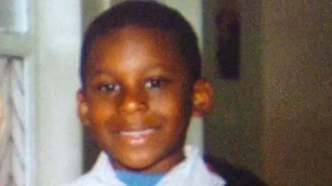 Family handout Anthony Walker, aged 10, is smiling at the camera pictured in the family home. He is wearing a white shirt with a blue top underneath.