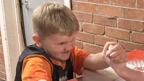 A young boy with blonde hair, wearing an orange and black t-shirt, and taking part in an arm wrestling competition. He has a look of determination on his face.