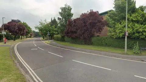 A residential road in Oxford, with a green hedge on the right and a red post box on the left. Trees line both sides of the road in the distance.