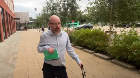 Chris Whitaker, with short fair hair and a close cropped beard, wearing rectangular steel-rimmed glasses, and a light blue shirt, is walking down a pavement with shrubs in the background. He is carrying a notebook in his right hand and using a walking stick with his left hand. 