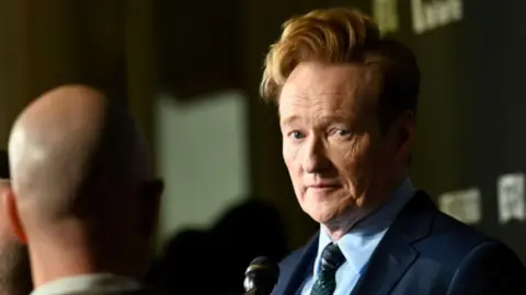 Conan O'Brien looks across the frame wearing a navy blue suit and blue tie.