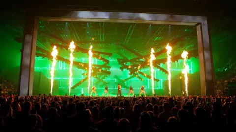 Reuters View of Bosnjak's performance from the back of the arena with the audience in the foreground. The stage is lit green and six tall flames are shooting into the air. Bosnjak is at the centre of the stage and is flanked by four female dancers.