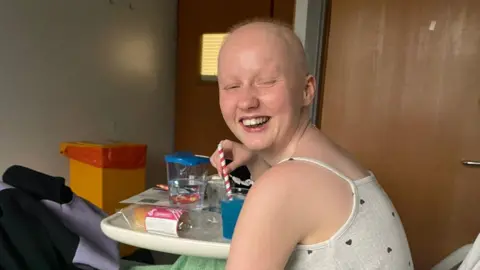 Enlli Thompson Enlli Thompson is photographed in a hospital bed. She is wearing a grey vest top which is covered in a pattern of small black hearts. She is drinking a blue slush drink and holding a white and red straw. She looks at the camera, smiling and winking.