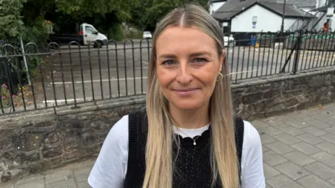 Beth is pictured wearing a black top and a white t-shirt underneath. She is looking at the camera and smiling, and behind her is a car park.