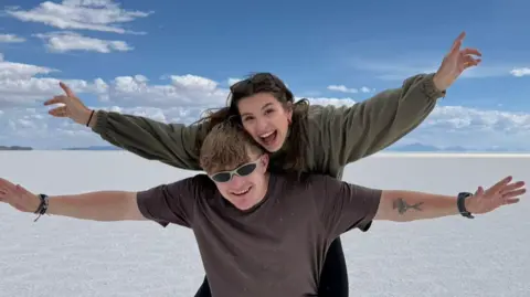 Sioned is pictured on Fin's back. They are both waving their arms in the air. They are pictured at the Uyuni Salt Flats in Bolivia. Fin is wearing sunglasses and they are both smiling.