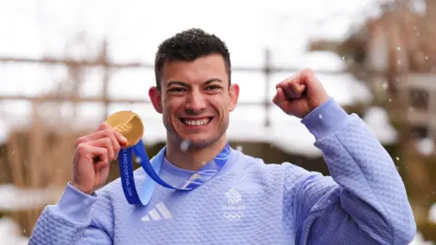 PA Media Great Britain's Skeleton gold medalist Matt Weston in Cortina d’Ampezzo, on day eight of the Milano Cortina 2026 Winter Olympics, Italy. He holds up his gold medal in one hand and punches the air with the other. He is wearing a light blue Team GB sweatshirt.