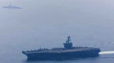 US Navy / Reuters The USS Abraham Lincoln sails at the bottom of the image on 6 February 2026. It is a large aircraft carrier with a flat runway. A number of military aircraft are stationary at the far side of the ship. The much smaller USS Frank E Petersen Jr is sailing at the top of the image. Source: US Navy / Mass Communication Specialist 1st Class Jesse Monford / Handout via Reuters