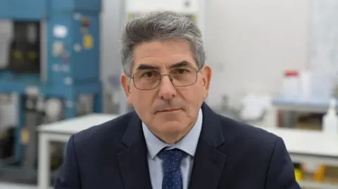 A man with grey hair is wearing a light blue shirt and dark-coloured suit jacket with a blue spotty tie. He is in what appears to be a factory. The room is brightly lit, and there are work benches and machinery behind the man.