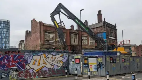 LDRS A digger disassembles a large red brick building behind metal screens. The building already has part of its roof and windows missing.