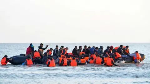 PA Media A group of people thought to be migrants wade through the sea wearing orange jackets to clamber aboard a small boat off the beach in Gravelines, France. 