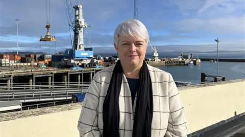 Hannah Beacom standing in front of Guernsey's harbour. She is wearing a cream coat with a black pattern, black top and black scarf. Her hair is white in a pixie cut and she is smirking. In the background there is an industrial site of crates and a crane. 